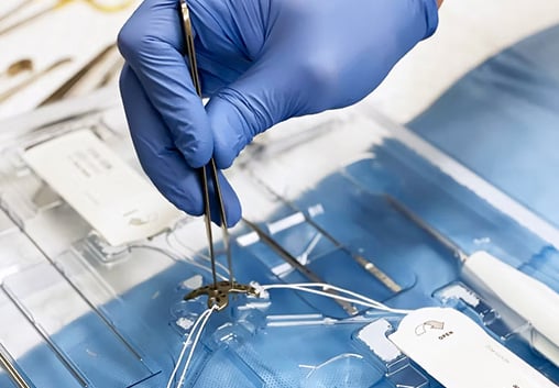 Gloved hands using forceps to lift a suture from an open sterile procedure kit