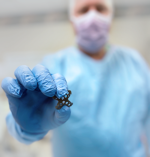 Surgeon in a mask and gown holding up the Endeavor The Summit™ Patella Plate between gloved fingers